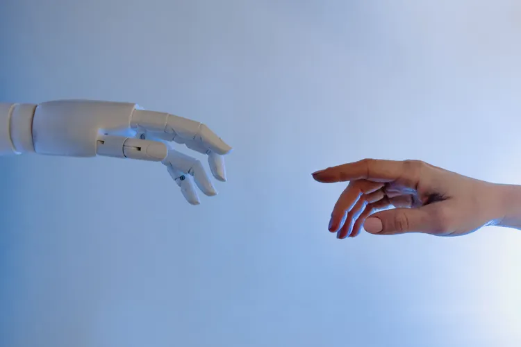 Een robotarm en een menselijke hand reiken naar elkaar. A.I neemt taken over, het werk zelf blijft deels mensenwerk.